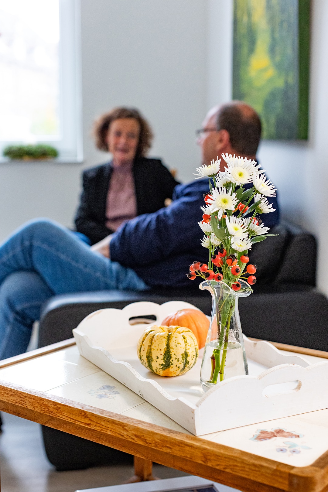 Blumenarrangement auf einem Tisch, während zwei Menschen in herzlichem Gespräch im Hintergrund sitzen, symbolisiert Trost.